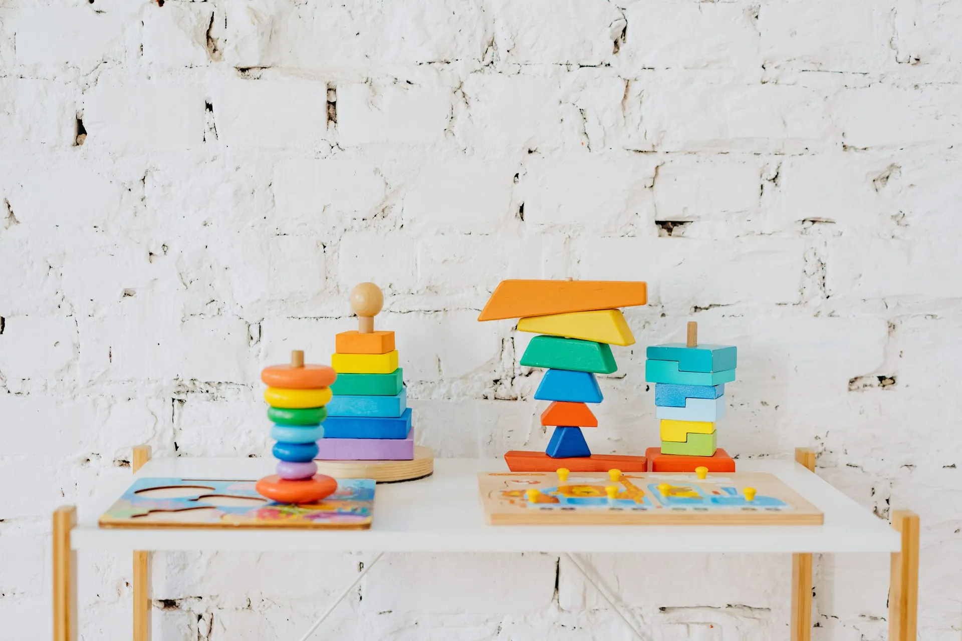 Colorful wooden stacking toys and peg puzzles on a small white table against brick