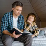 Father reading a hardback book aloud to his son on a cozy attic bed