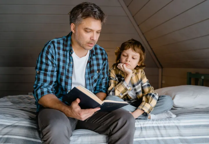 Father reading a hardback book aloud to his son on a cozy attic bed