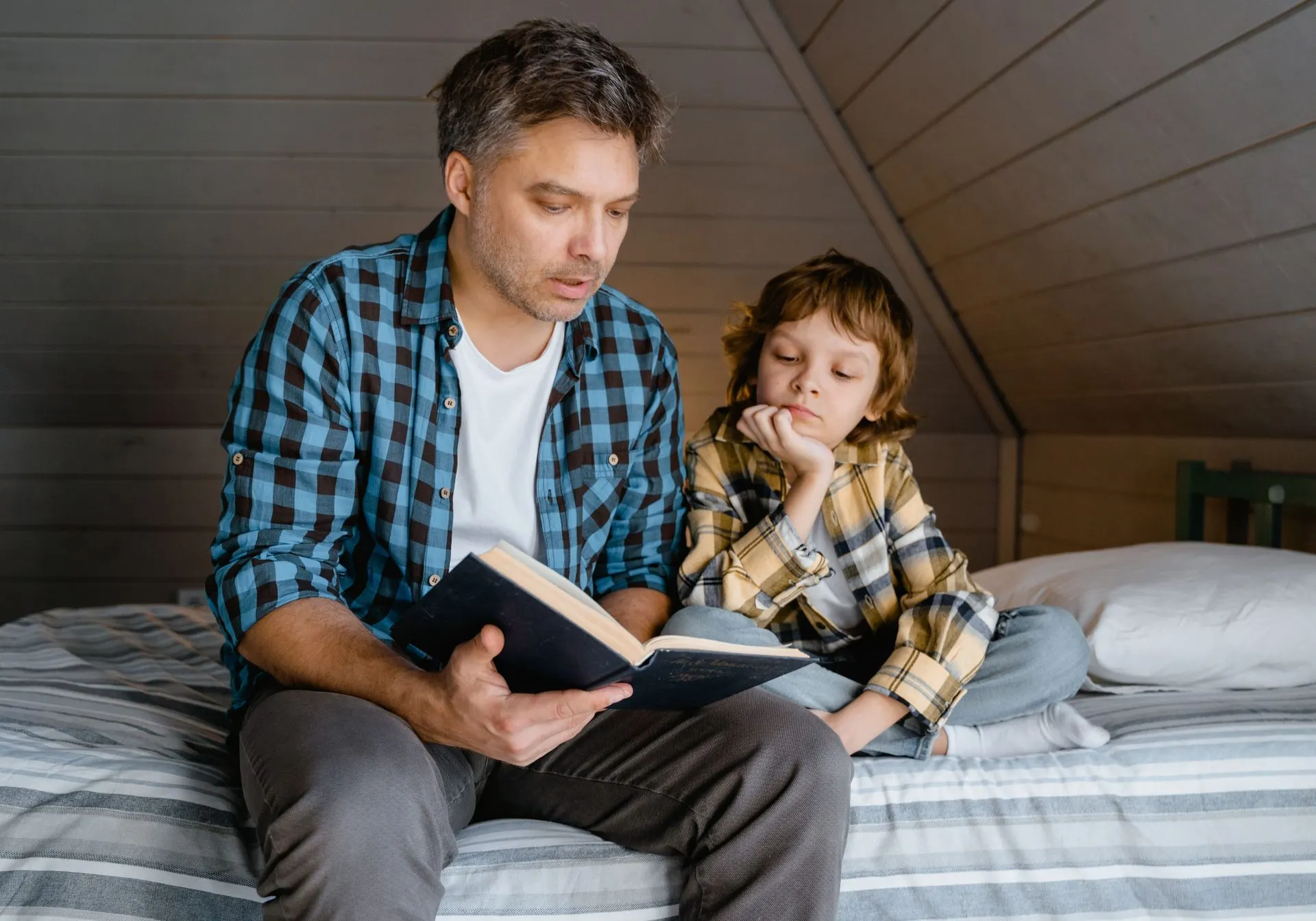 Father reading a hardback book aloud to his son on a cozy attic bed