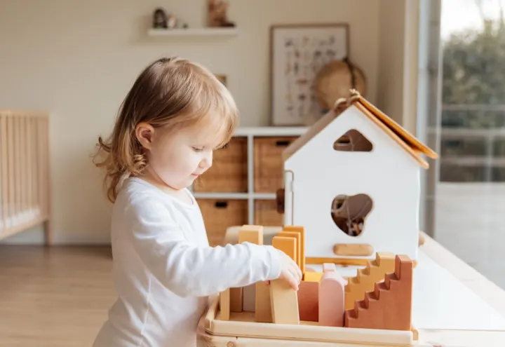 Little girl arranging wooden blocks beside a white toy house in a sunny playroom
