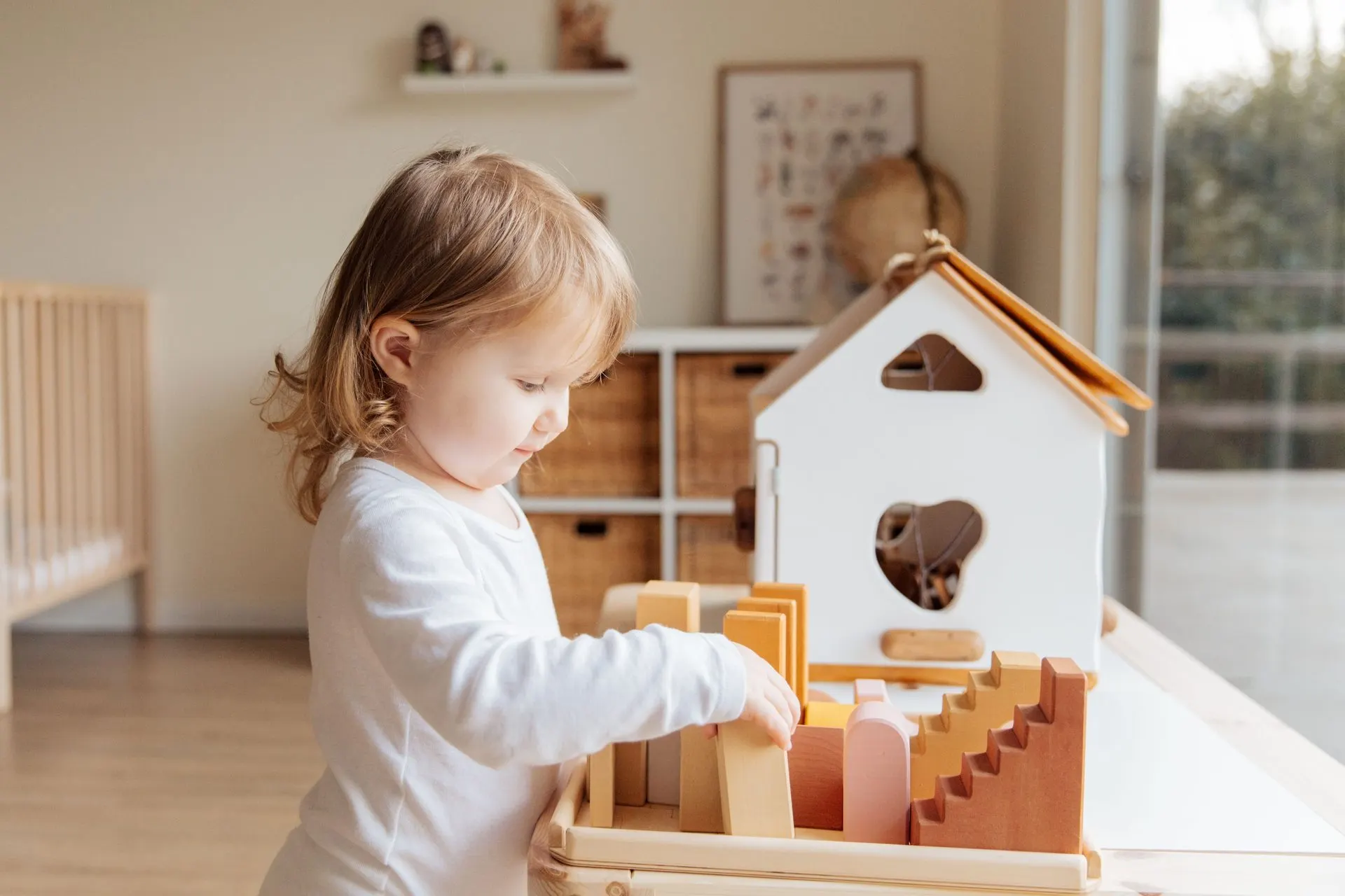 Little girl arranging wooden blocks beside a white toy house in a sunny playroom