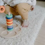Toddler adding an orange ring to a rainbow wooden stacker on a fluffy rug