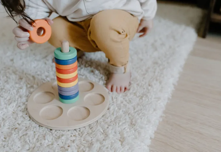 Toddler adding an orange ring to a rainbow wooden stacker on a fluffy rug