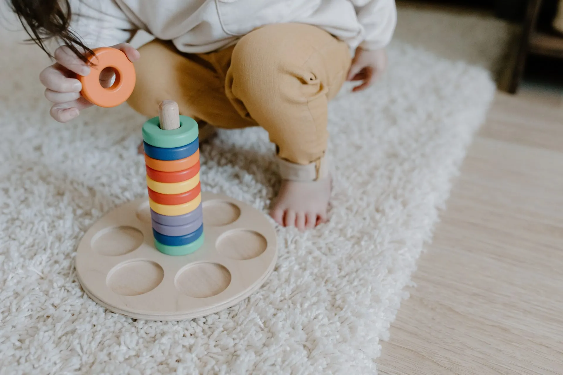 Toddler adding an orange ring to a rainbow wooden stacker on a fluffy rug
