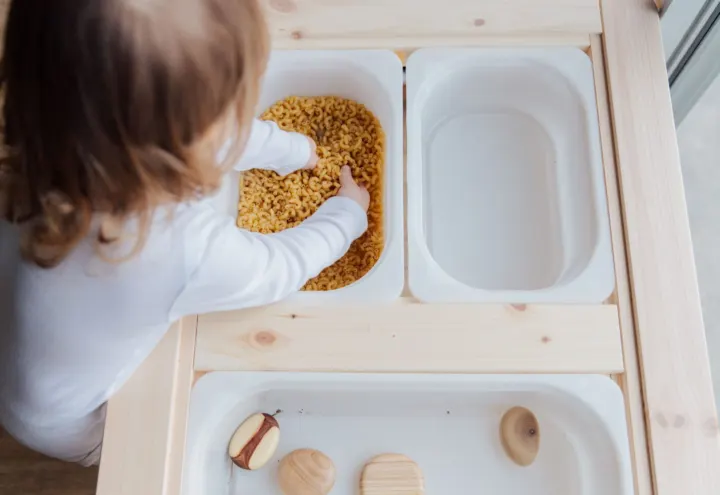 Toddler scooping dry pasta in a white sensory bin on a wooden play table