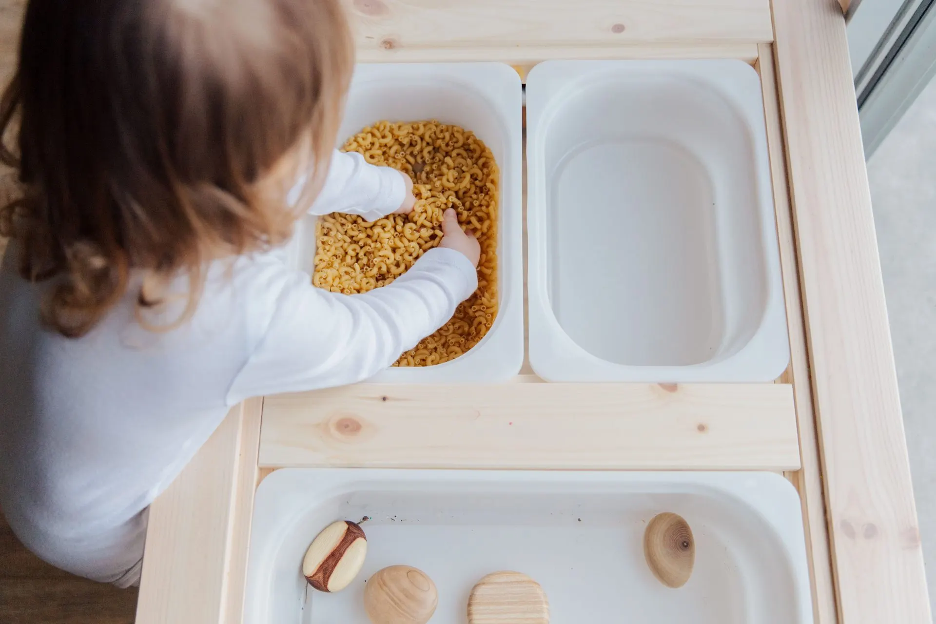 Toddler scooping dry pasta in a white sensory bin on a wooden play table