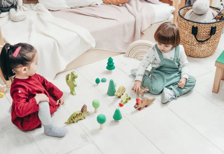 Two young children playing with wooden dinosaurs and trees on a bright bedroom floor