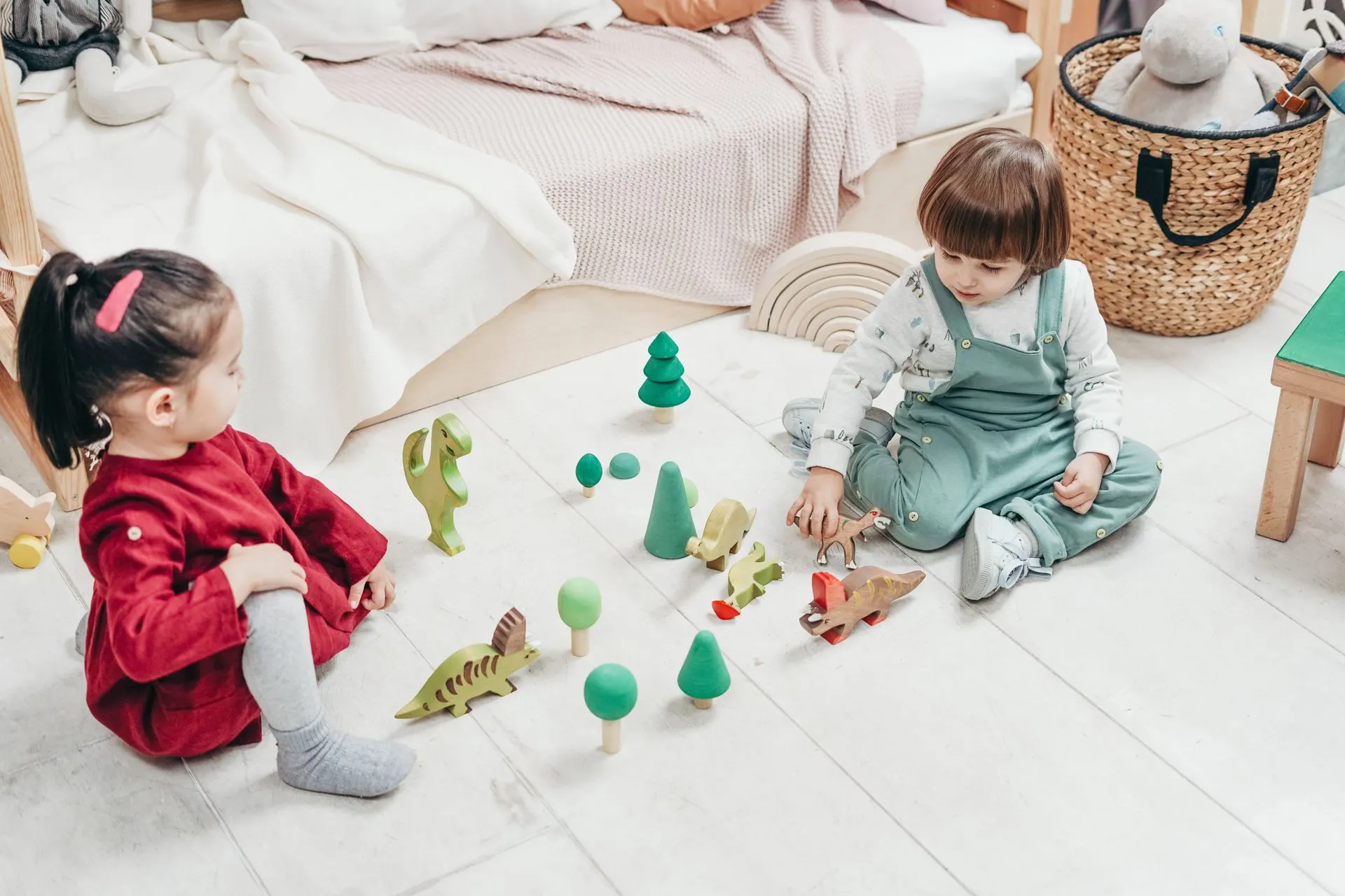 Two young children playing with wooden dinosaurs and trees on a bright bedroom floor